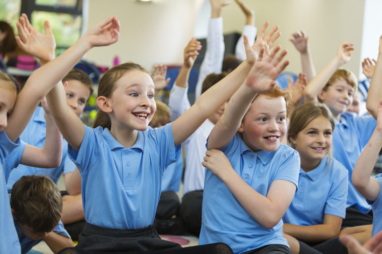 Excited school children in school uniform with hands up ready to answer a question from the teacher