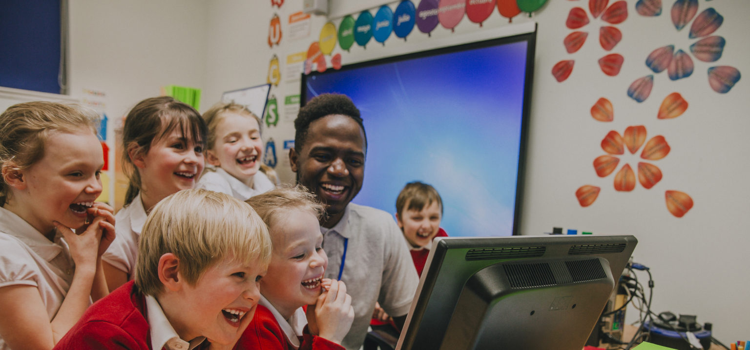 Group of primary school students are crowded round a computer with their teacher. They are all laughing and looking at the computer screen.